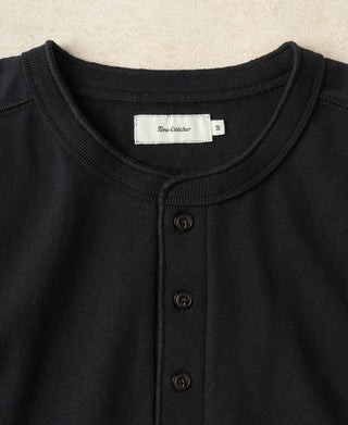 Close-up of a black henley shirt with a ribbed crew neck and three dark buttons, featuring a white neck label that reads 'Time Catcher'.