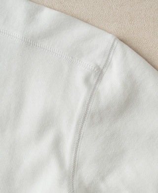 Close-up of a white ribbed knit fabric, showing the texture and a neat hemstitch detail against a neutral beige background.