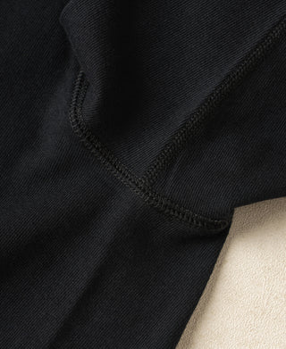 Close-up of a black ribbed knit garment showing a folded corner and contrast stitching, with a small area of light beige fabric visible at the lower right.